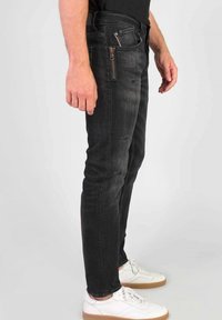 Hombre vistiendo jeans negros ajustados con un bolsillo con cremallera lateral y zapatillas blancas, de pie contra un fondo gris claro.