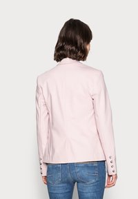 Femme aux cheveux bruns courts et ondulés portant un blazer rose clair et un jean bleu, vue de dos sur un fond clair uni.