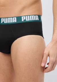 Κοντινό πλάνο άνδρα που φοράει μαύρα εσώρουχα με πράσινη μέση με λευκό λογότυπο «PUMA», δείχνοντας τους γοφούς και το δεξί χέρι χαλαρό στο πλάι.