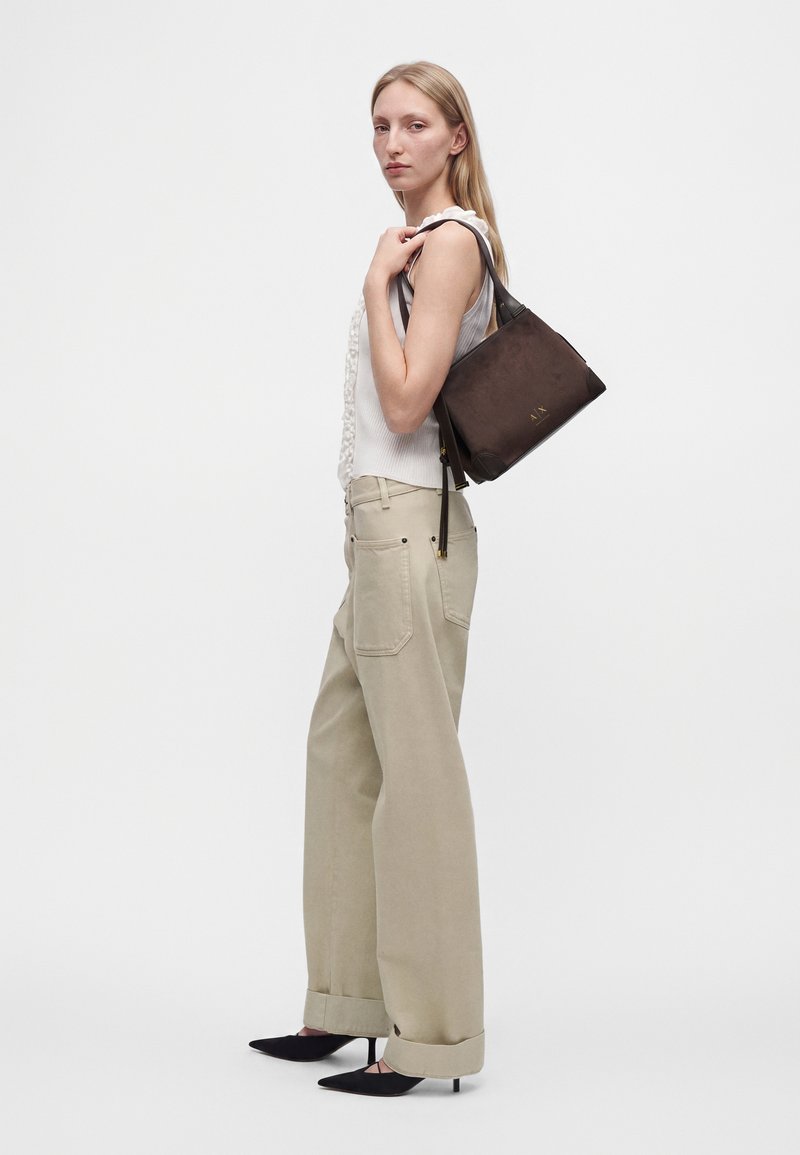 Femme portant un pantalon beige à jambes larges et un top blanc sans manches, tenant un sac bandoulière marron, debout devant un fond clair uni.