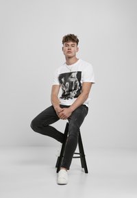 White t-shirt featuring a black and white graphic print, paired with distressed black jeans and white sneakers, seated on a black stool.