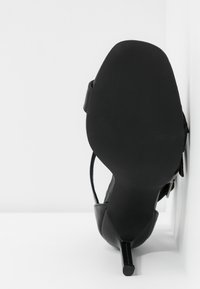 Sandale à talon haut noire avec un bout pointu et un talon fin, vue de la semelle face à l'appareil photo sur un fond blanc.