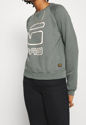 Osoba ubrana w zieloną bluzę G-Star Raw z logo z przodu, czarne spodnie i złoty łańcuszek na szyi, stojąca na białym tle.