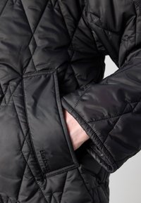 Zwarte gewatteerde jas met een ruitpatroon, een gladde textuur en een zichtbare zak aan de voorkant. Barbour-logo vlakbij de zak.