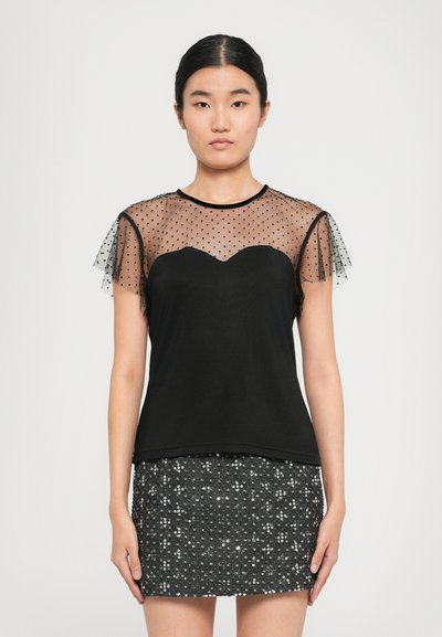 Femme portant un haut noir avec des manches courtes transparentes à pois et un décolleté en cœur, associé à une mini-jupe noire pailletée.