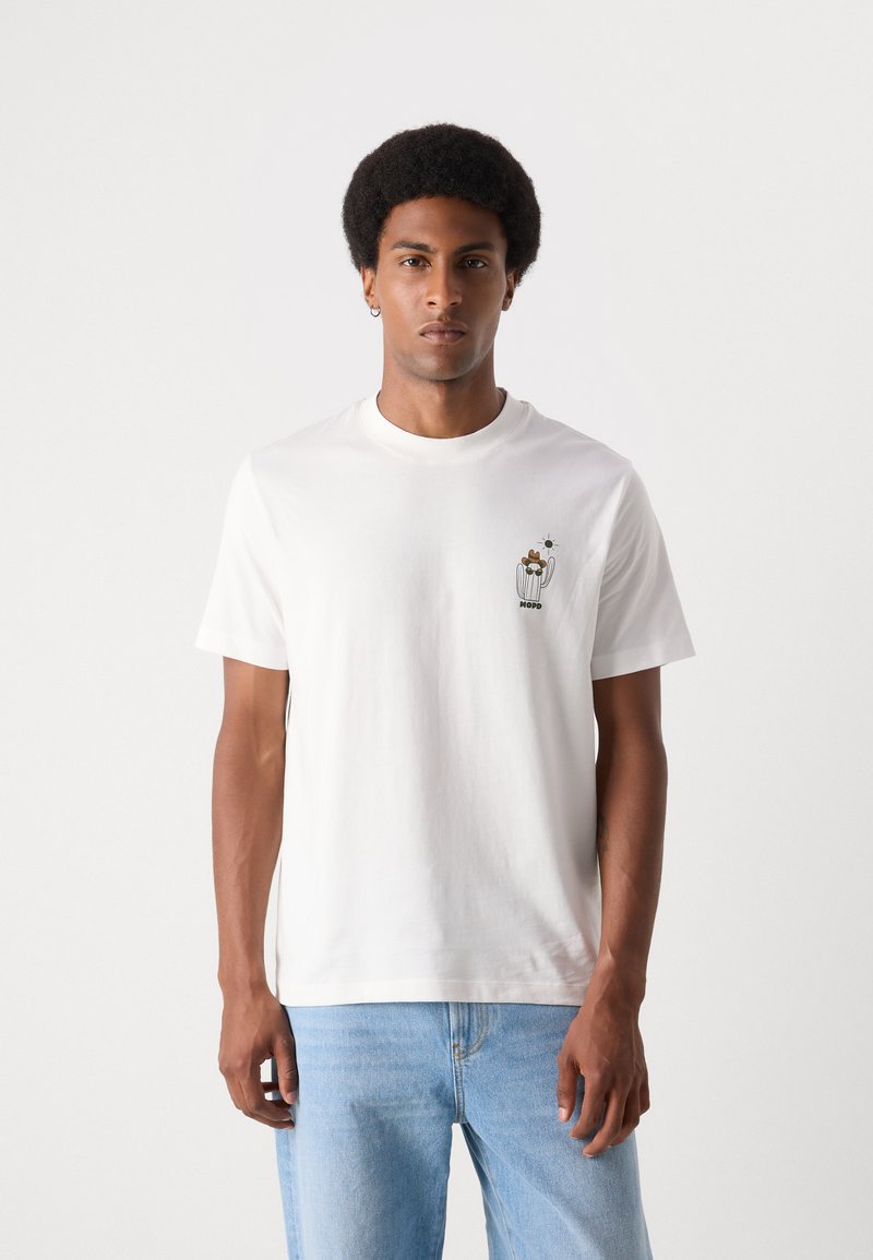 Weißes Baumwoll-T-Shirt mit kurzen Ärmeln, das auf der linken Brust ein kleines grafisches Motiv in Braun- und Grüntönen zeigt, präsentiert von einem Modell.