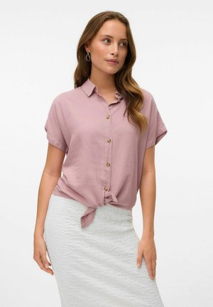 Blusa rosa chiaro a maniche corte con chiusura a bottoni e dettaglio annodato sull'orlo, abbinata a una gonna bianca strutturata.