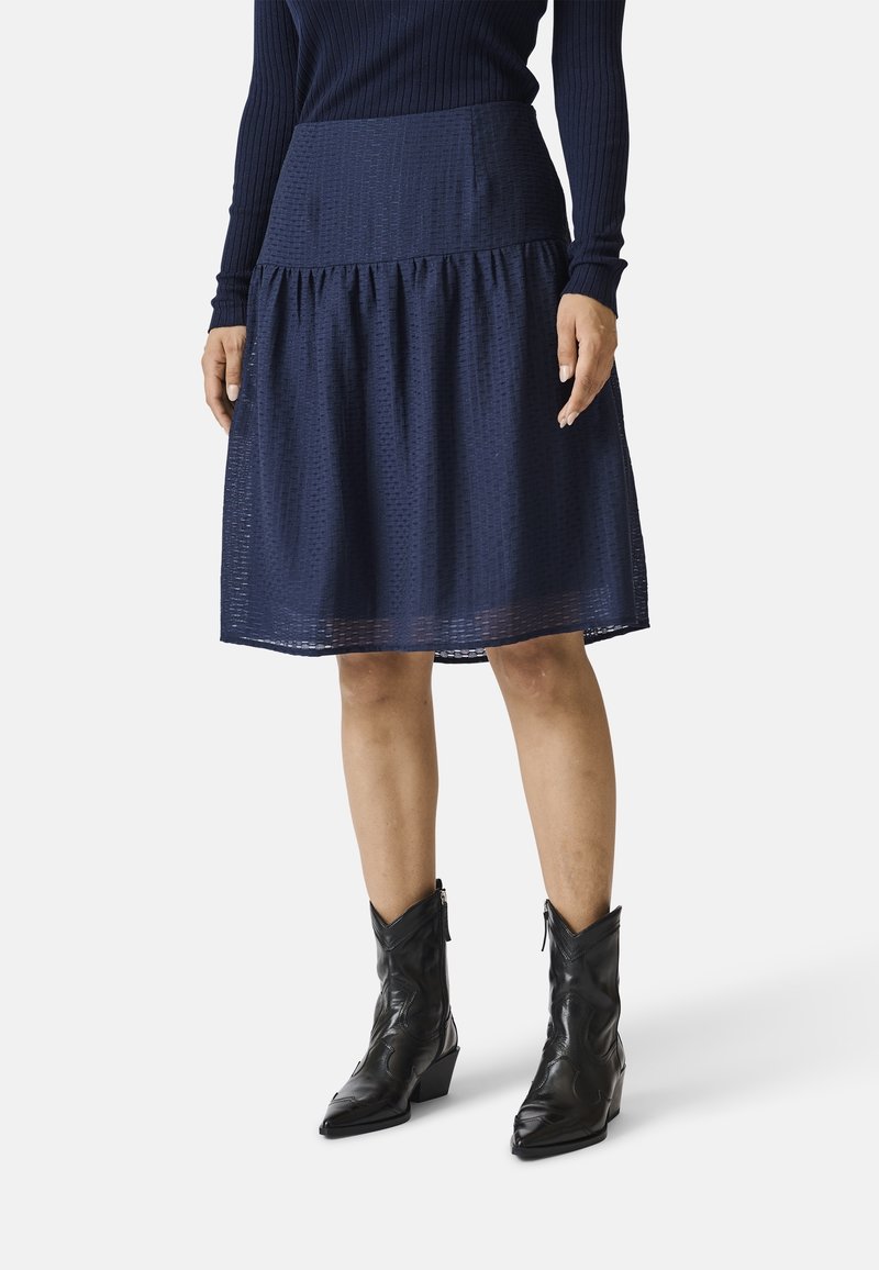 Femme portant une jupe texturée bleu marine longueur genou, des bottes de cowboy noires à bout pointu, et un haut à manches longues côtelé bleu marine.