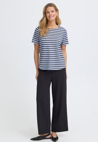 Mujer sonriendo de pie, vestida con una camiseta de manga corta a rayas azules y negras, pantalones negros de pierna ancha y zapatos planos negros con punta afilada.