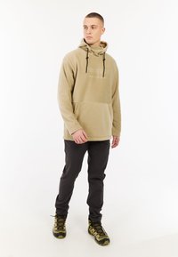 Beige fleecetrui met een trekkoord, kangoeroezak en reliëflogo; gecombineerd met donkere broek en geblokte wandelschoenen.