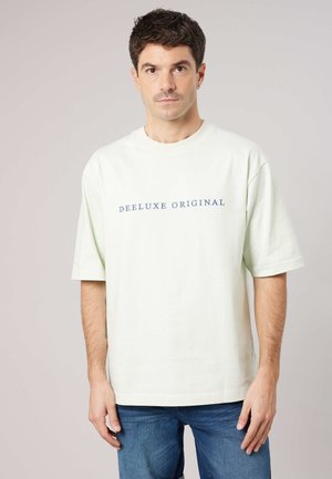 Man met kort donker haar, draagt een wit oversized T-shirt met de tekst "DEELUXE ORIGINAL" en een blauwe spijkerbroek, staand tegen een lichte achtergrond.