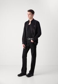 Camisa de mezclilla negra superpuesta sobre una camiseta negra sin mangas, combinada con jeans negros y zapatos negros. Diseño simple, sin patrones prominentes.