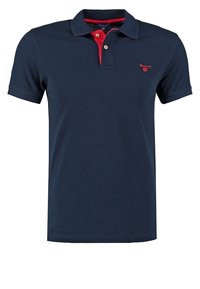Polo bleu marine à manches courtes avec col intérieur rouge, deux boutons et petit logo GANT rouge sur la poitrine gauche.