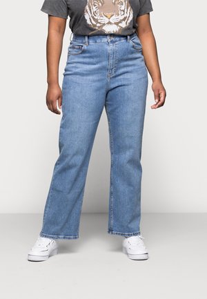 Person iført lyseblå jeans med lige ben, hvide sneakers og en mørk t-shirt med et tigeransigtsgrafik.