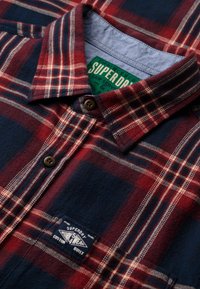 Chemise en flanelle à carreaux rouge et marine avec un col boutonné, des boutons marron et une patch cousue arborant les détails de la marque en bas à gauche.