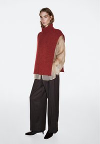 Frau trägt einen roten ärmellosen Strickrollkragenpullover über einer transparenten beigen Bluse, weite dunkle Hosen und schwarze spitze Schuhe, steht vor weißem Hintergrund.