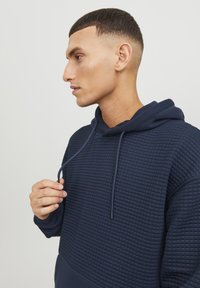 Dunkelblauer Kapuzenpullover mit strukturiertem Oberbereich und glattem Unterteil, ausgestattet mit einer Kordel und einem lässigen Schnitt.