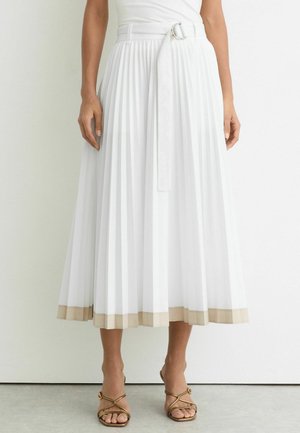 Kvinde iført hvid plisseret midi-nederdel med beige kant og bælte, kombineret med beige rem-sandaler med åben tå og hæl.