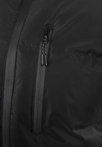 Giacca nera isolante con tasca con zip, tessuto liscio e cuciture visibili. La cerniera è dotata di una linguetta per una facile apertura.