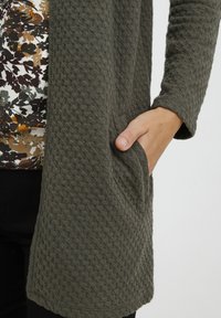 Olivgrön strukturerad kofta med ett vadderat mönster, med långa ärmar och en framficka. Lager på en blommönstrad topp.