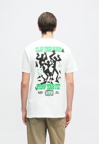 Λευκό βαμβακερό μπλουζάκι με έναν μαύρο και πράσινο γραφικό σχεδιασμό στην πλάτη, ο οποίος περιλαμβάνει κείμενο "ΧΤΥΠΗΣΕ ΤΑ ΧΕΡΙΑ ΣΟΥ" και "ΓΔΥΣΕ ΤΑ ΠΟΔΙΑ ΣΟΥ." Με κοντά μανίκια.