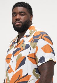 Camisa de manga corta con una base blanca y grandes patrones abstractos de hojas en naranja, azul marino y verde. Tela ligera y texturizada.