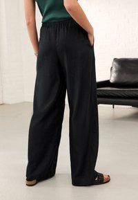 Svarte bukser med vide ben og snøre i livet, lavet af et let stof. Sidelommer og løs pasform er synlige. Sandaler er båret.