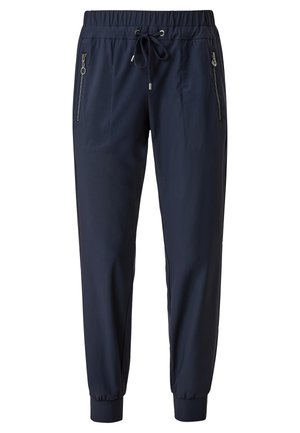Persona que lleva una camiseta blanca de manga corta, pantalones jogger azul marino con bolsillos con cremallera y cordón, y zapatillas blancas con cordones, de pie sobre un suelo beige.