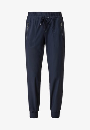Personne portant une chemise blanche à manches courtes, un pantalon de jogging bleu marine avec poches zippées et cordon, et des baskets blanches à lacets, debout sur un sol beige.