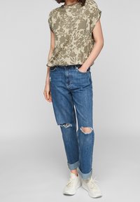 Beżowa bluzka z krótkim rękawem w kwiatowy wzór, zestawiona z przetartymi niebieskimi dżinsami i białymi trampkami. Dżinsy podwinięte u mankietów.