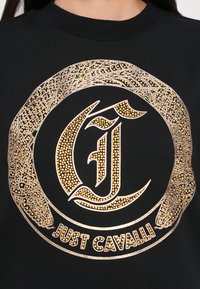 Schwarzer Pullover mit einem strukturierten goldenen Design, das ein kreisförmiges Motiv sowie ein markantes "C" und den Text "JUST CAVALLI" darunter zeigt.
