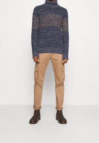 Pull à col roulé en laine bleu marine avec des rayures horizontales beiges, accompagné d'un pantalon cargo beige. Chaussures : bottines marron foncé.