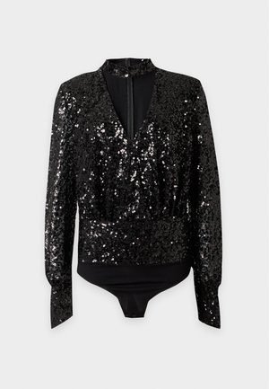 Femme aux cheveux bouclés et foncés porte un chemisier noir à sequins avec un détail col ras-du-cou et un pantalon noir à jambes larges, debout devant un fond clair.