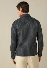 Muška prugasta košulja od tamnog materijala s okomitim bijelim linijama, s širokim ovratnikom i dugim rukavima. Pogled sa stražnje strane prikazuje strukirani kroj.