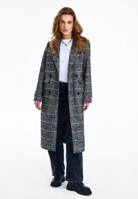 Žena s vlnitými vlasmi, oblečená v dlhej sivej kockovanej bunde, bielom topu, čiernych širokých nohaviciach, čiernych robustných topánkach a náhrdelníku, stojaca na bielom pozadí.