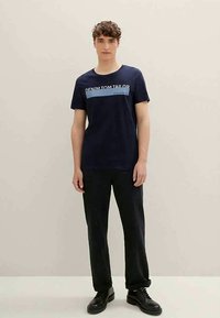 Navy cotton t-shirt with short sleeves, featuring a white and light blue graphic text "DENIM TOM TAILOR." Worn with black pants and shoes.