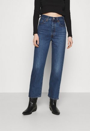 Personne portant un jean bleu taille haute à jambes droites, associé à des bottines noires et un top noir cropped à manches longues, sur un fond uni.
