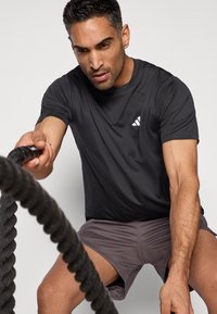 Svart atletisk t-skjorte med korte ermer og en liten hvit logo, kombinert med grå teksturerte shorts. Personen holder et tykt, svart tau.