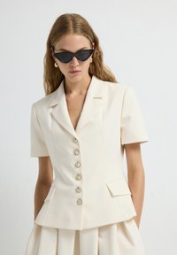 Donna che indossa un blazer cortissimo color crema con bottoni perlacei e una gonna abbinata, accessoriata con occhiali da sole neri a forma di cat-eye e orecchini d'oro.