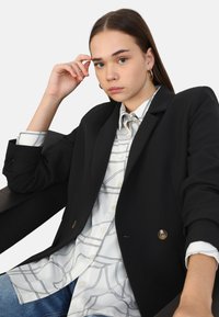 Blazer negro ajustado con botones dorados sobre una camisa blanca con un patrón de rejilla negra, combinado con jeans de mezclilla azul claro. Textura de tela suave.