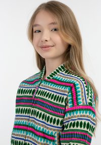 Többszínű mintázatú zipzáras dzseki, zöld, rózsaszín, kék és arany kiegészítőkkel; texturált anyagból készült, levelekre emlékeztető és geometrikus mintákkal.