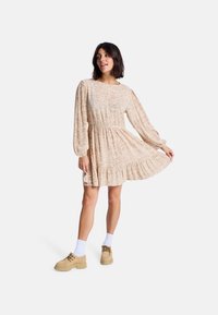 Robe beige à manches longues et jupe à volants avec un motif de feuilles, cintrée à la taille. Associée à des chaussures beiges et des chaussettes blanches.