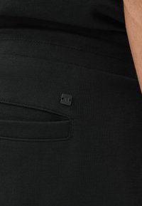 Pantalones jogger de tela negra con una pequeña etiqueta cuadrada negra y un bolsillo lateral. El material parece suave con una textura lisa.