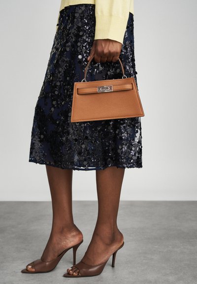Bolso de mano texturizado marrón con un diseño estructurado y un cierre plateado, sostenido en la mano de una mujer, combinado con una falda de lentejuelas azul marino y sandalias de tacón marrones.