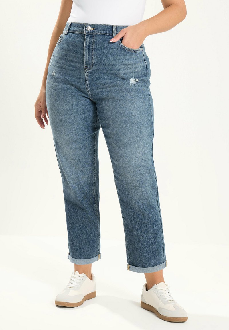 Kobieta w podwiniętych niebieskich dżinsach z delikatnymi przetarciami i białych tenisówkach, z ręką w kieszeni na białym tle.