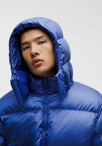 Sinine polsterdatud jakk kõrge kraega ja suure kapuutsiga. Varustatud lukuga ja läikiva tekstuuriga. Pole nähtavaid mustreid ega aktsente.