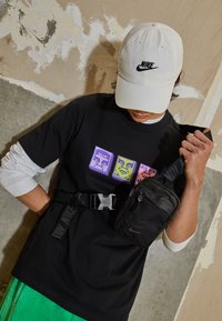 Svart t-shirt med färgglada grafiska rutor, gröna shorts, svart crossbody-väska och vit keps med svart logotyp. Texturerad bakgrund.