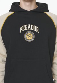 Felpa con cappuccio nera e beige con maniche raglan, caratterizzata dalla scritta "PEGADOR" e dal logo in oro, e una tasca a marsupio frontale.