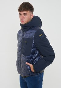 Marineblauwe pufferjack met een glanzend, gewatteerd ontwerp, voorzien van een capuchon, ritszakken en gele accenten. Gemaakt van lichtgewicht, geïsoleerd materiaal.