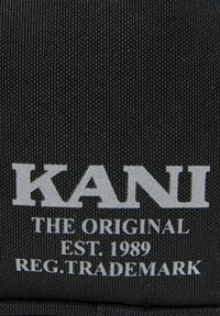 Sort tekstil med en tekstureret overflade. Har "KANI" i fede sølv store bogstaver, med "THE ORIGINAL EST. 1989" og "REG. TRADEMARK" nedenfor.